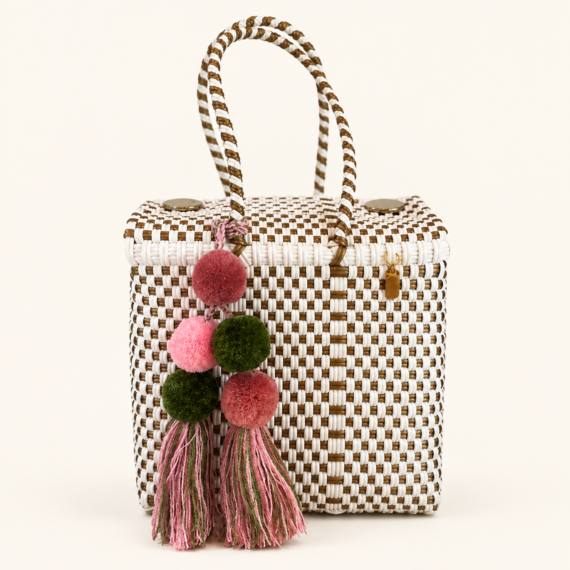 A woven straw tote bag with brown and beige checkered pattern, featuring dual top handles and a decorative hanging tassel with pink and green pom-poms, set against a warm beige background.