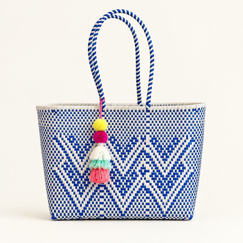 A woven tote bag with a blue and white geometric pattern, featuring two braided blue and white handles and a colorful tassel charm, set against a solid light beige background.