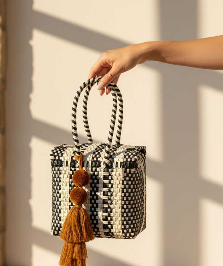 A woven handbag with black and off-white geometric stripes and a large mustard-yellow tassel, displayed on a round wooden stool against a plain light gray wall.