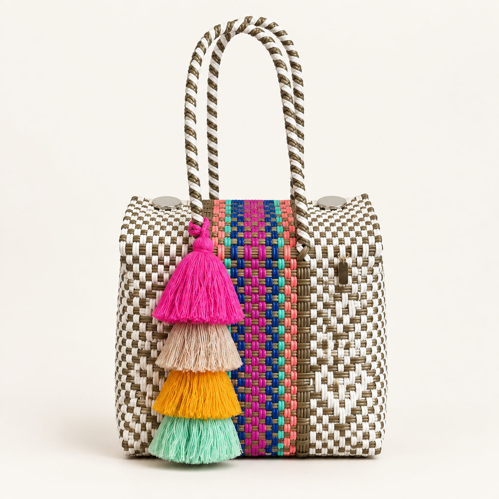 A woven tote bag with a cream and brown geometric pattern, a colorful striped accent panel, and a large multi-colored tassel, displayed on a wooden stool against a neutral wall.