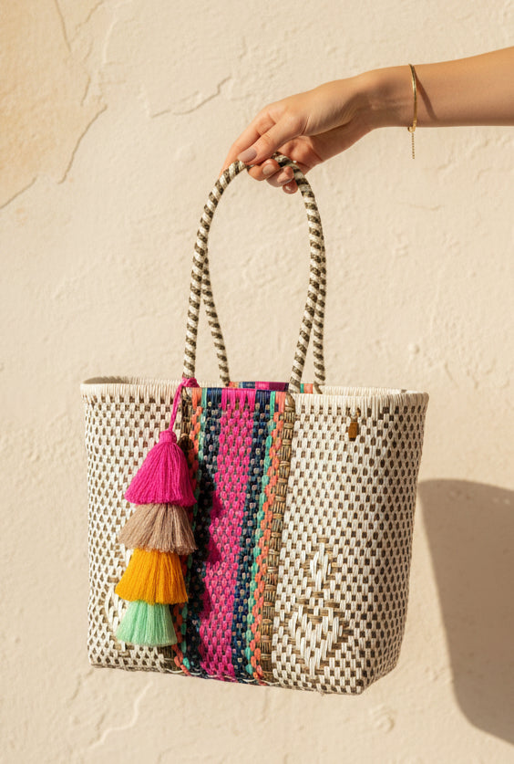 A woven tote bag with a cream and brown geometric pattern, featuring a vibrant pink, teal, and brown vertical stripe detail on the front and a tiered tassel charm in pink, beige, yellow, and mint green. The bag is displayed on a wooden stool against a neutral gray wall.