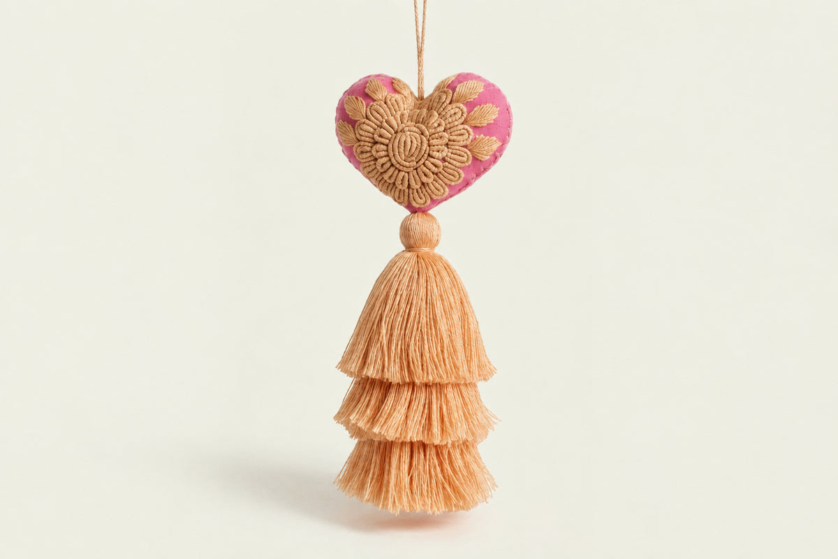 A decorative hanging ornament with a pink and beige embroidered heart on a string, above a tiered beige tassel, photographed against a soft cream background with blurred dried grass and a white vase.