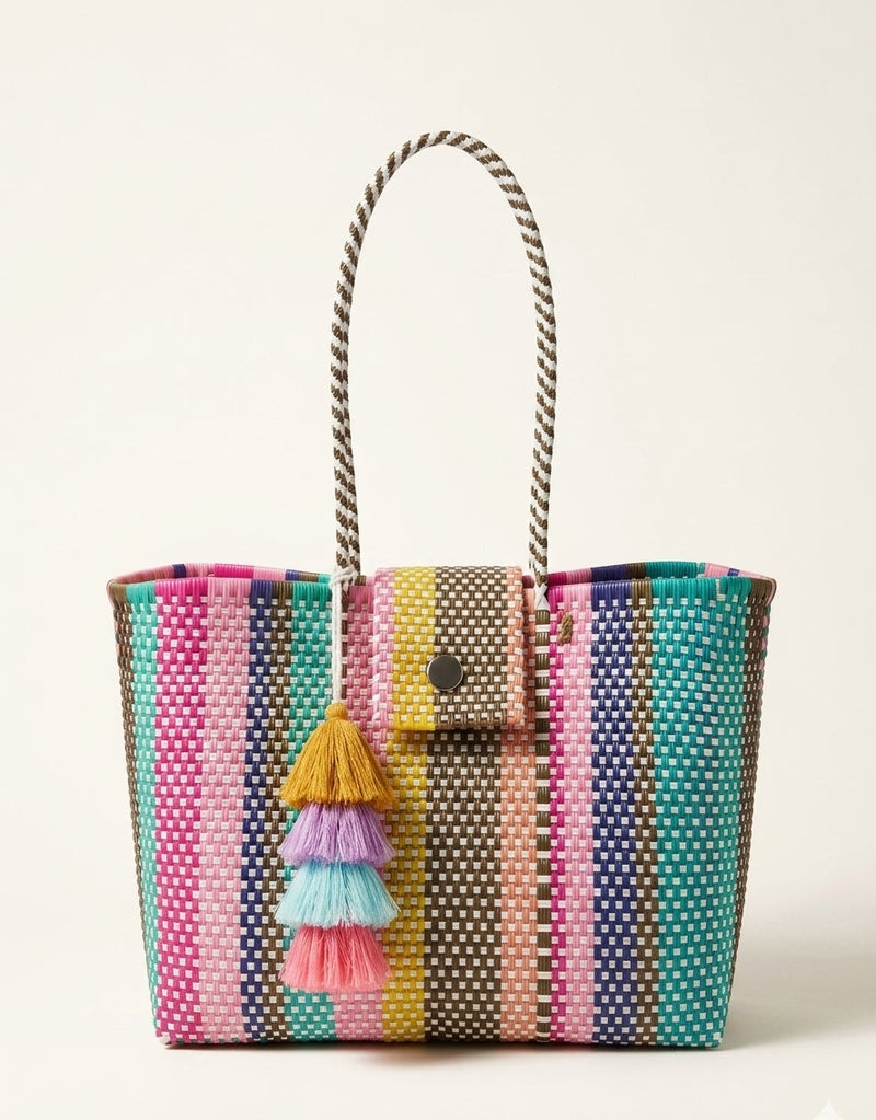 A woven tote bag with vertical stripes in pink, teal, yellow, brown, and navy, featuring a small front pocket with a tassel charm, set against a plain off-white background.